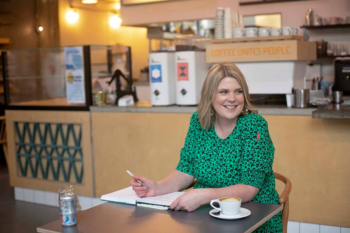 Hannah Brooke Photographer sitting in a cafe, writing in a notebook, smiling and looking to her left