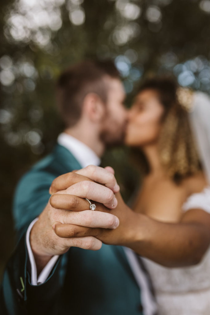 A close up of the ring with the bride and groom kissing in the background