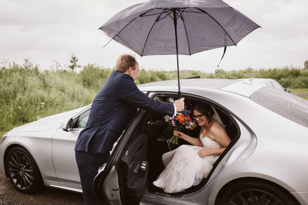 A groomsmen helps the excited bride out of the car in the rain