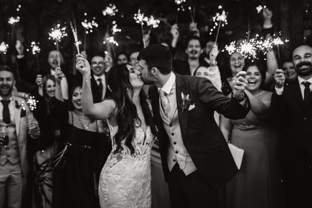 Bride & Groom kiss whilst holding sparklers with the wedding guests