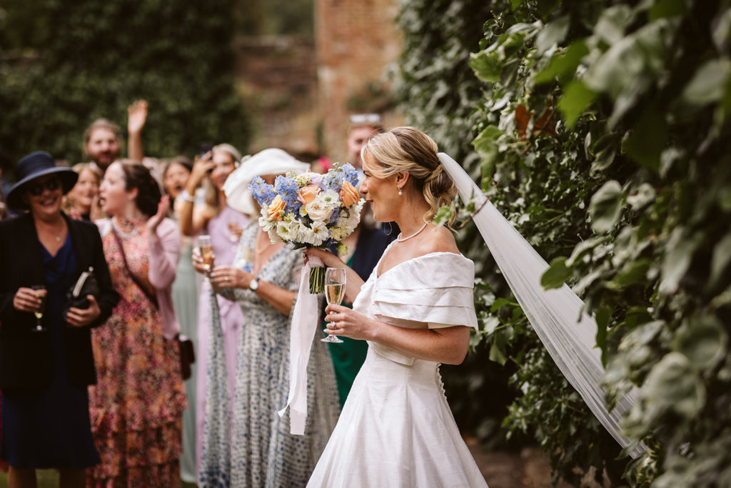 Bride enters her outdoor drinks reception as guests look on.