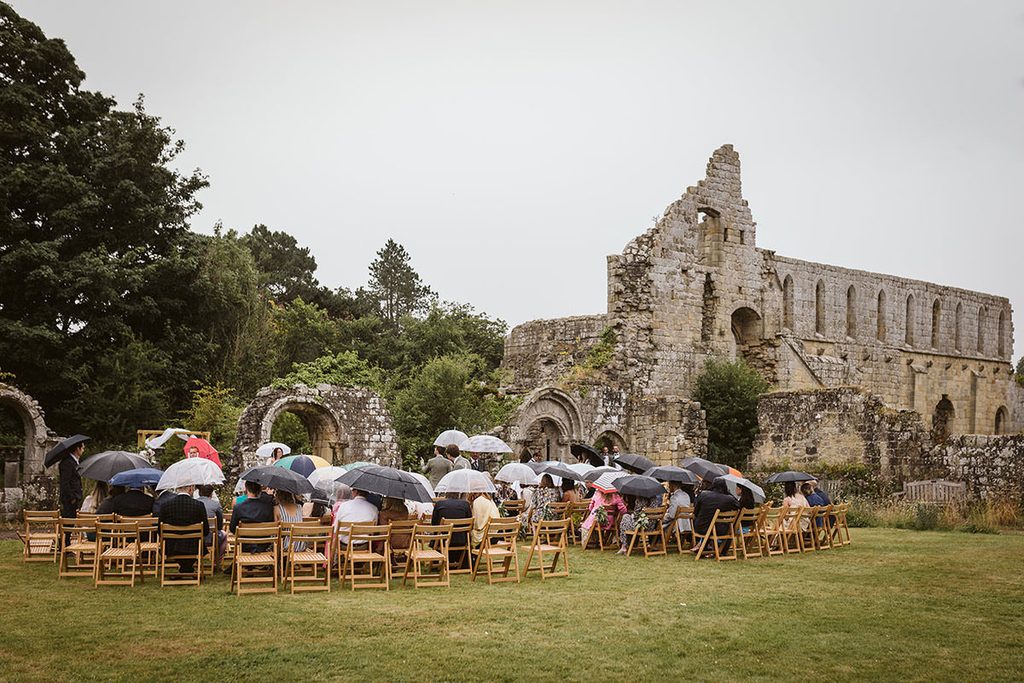 Guests sheltering under umbrellas during an outdoor ceremony at Jervaulx Abbey